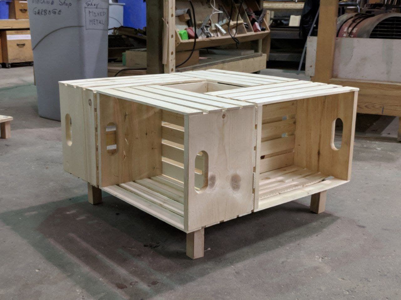 the table before staining. four box-like wine crates are on their sides and joined together in a spiral. a messy wood shop is in the background